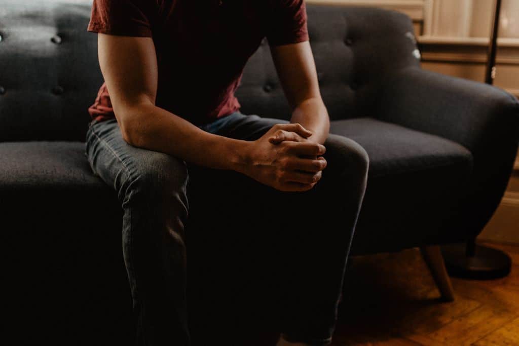 A close up of a man's body sitting on a grey Scandinavian sofa in a cocaine addiction therapy session with his hands clasped and elbows on knees - Which Rehab